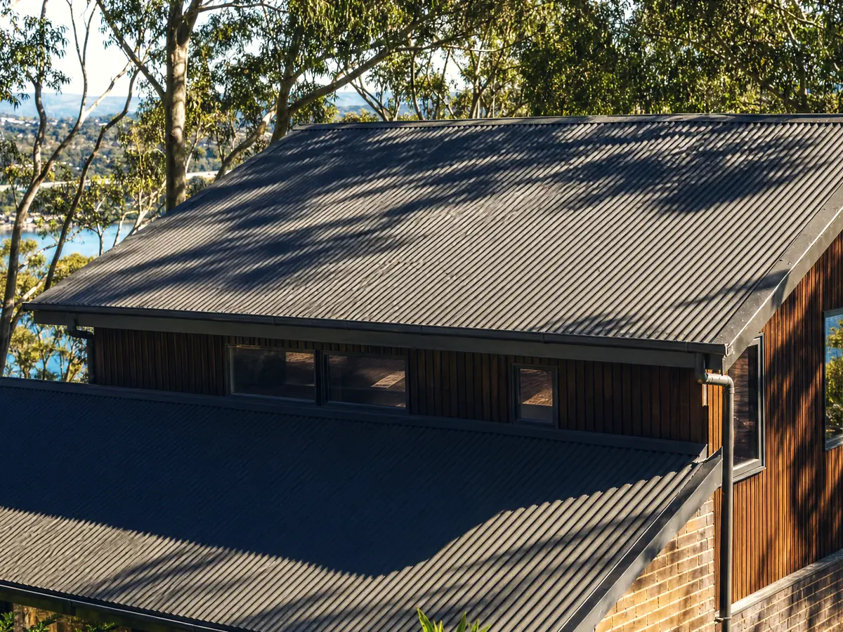 A split-level Toronto home on a steep block, its dark Colorbond roof dappled by the gum canopy overhead, lake water below