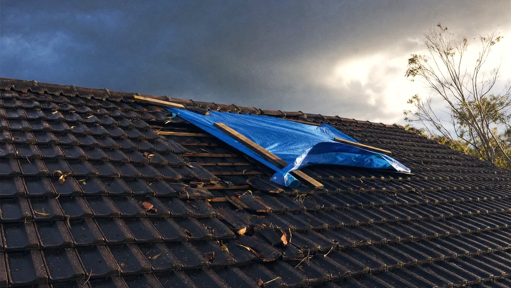 Tile roof after a storm with several tiles lifted and displaced, tarpaulin partly over the gap, wet sky behind